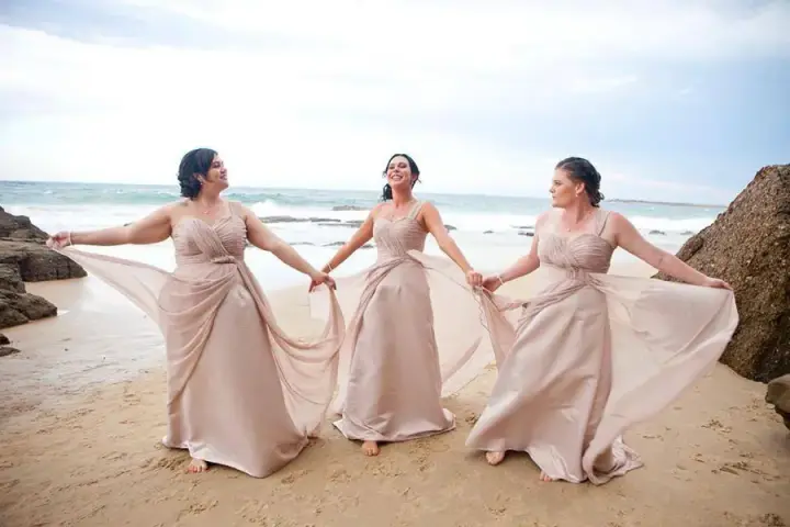 Three bridesmaids on the beach in blush satin gowns, each with her hair styled into an updo