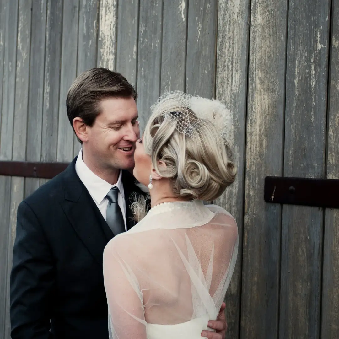 Bride in a birdcage veil with a vintage pinned style, kissing her groom against a timber wall
