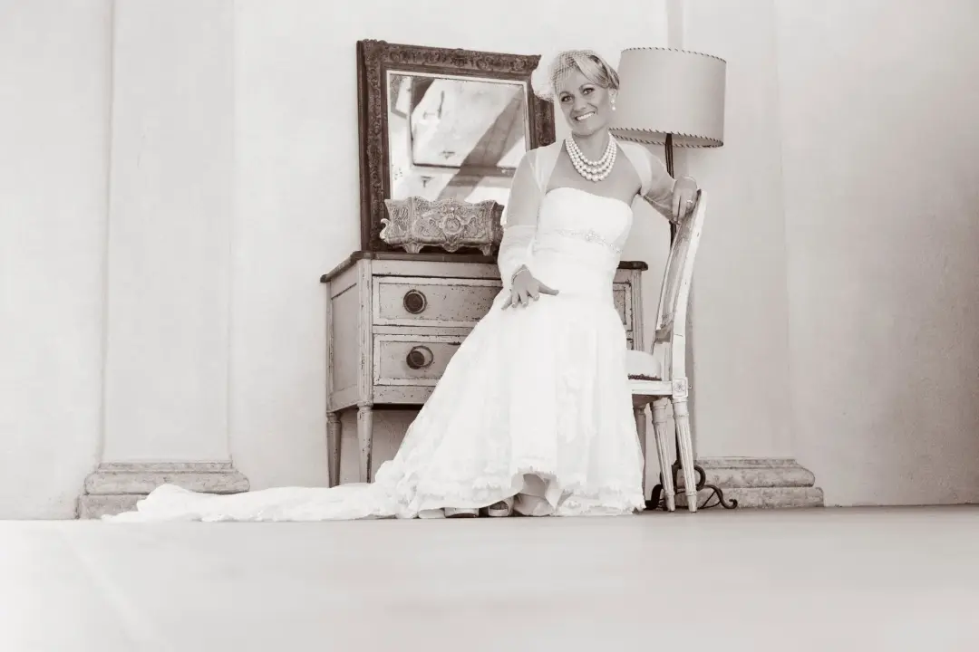 Bride seated at a dressing table in a tea length gown, her short blonde hair styled with a pearl necklace