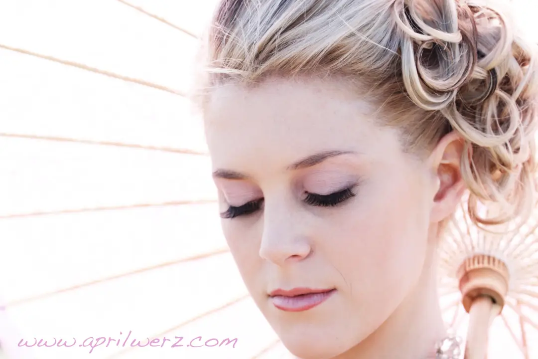 Bride under a parasol with her blonde hair curled into a pinned updo, wearing soft neutral makeup
