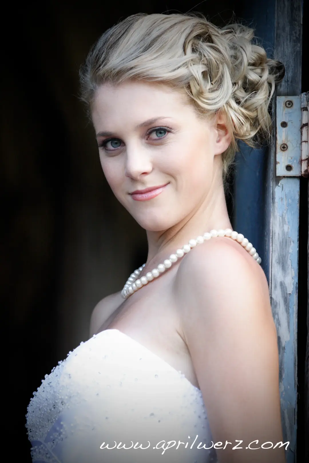 Bride against a timber door, her blonde hair curled into a pinned updo with a pearl strand