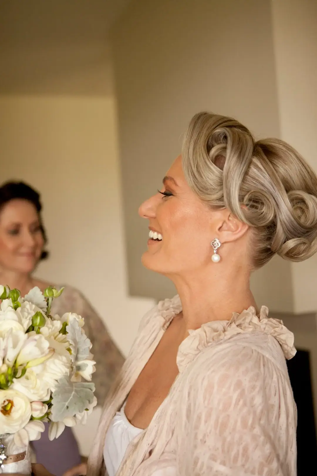 Mother of the bride in a rolled vintage updo with pearl drop earrings, holding a white bouquet