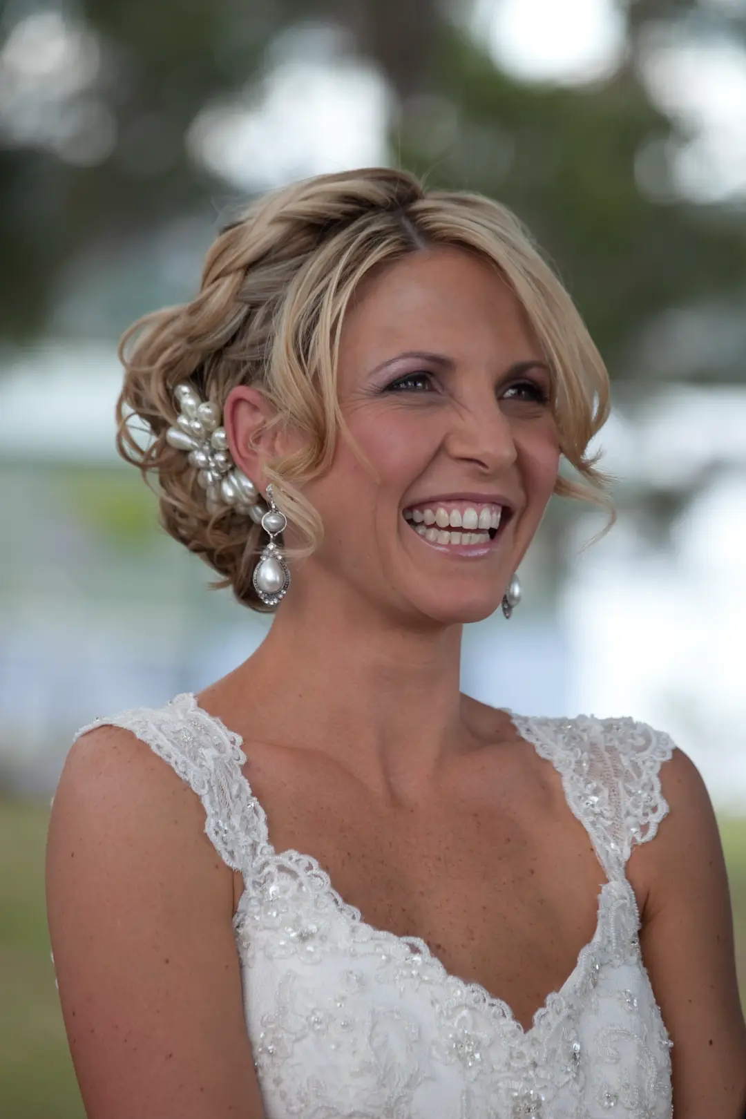 Bride in a curled blonde updo with a pearl hairpiece and matching drop earrings, wearing a beaded lace gown