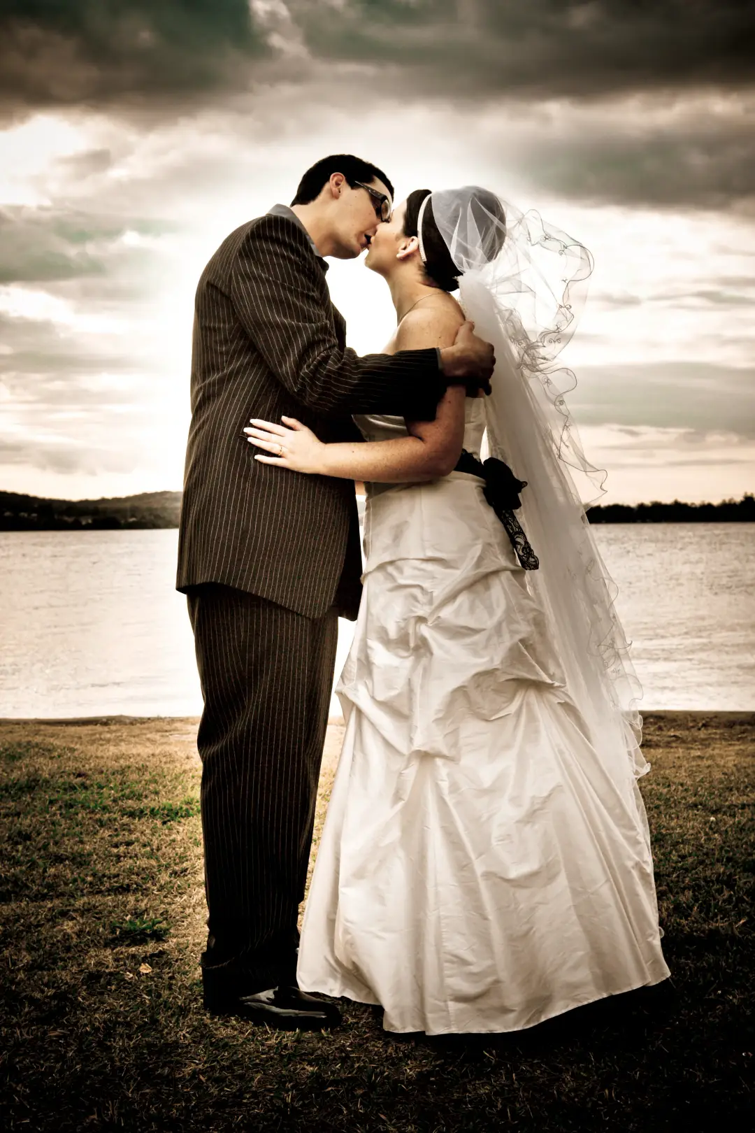 Bride and groom kissing by the lake, her veil pinned into a dark low updo