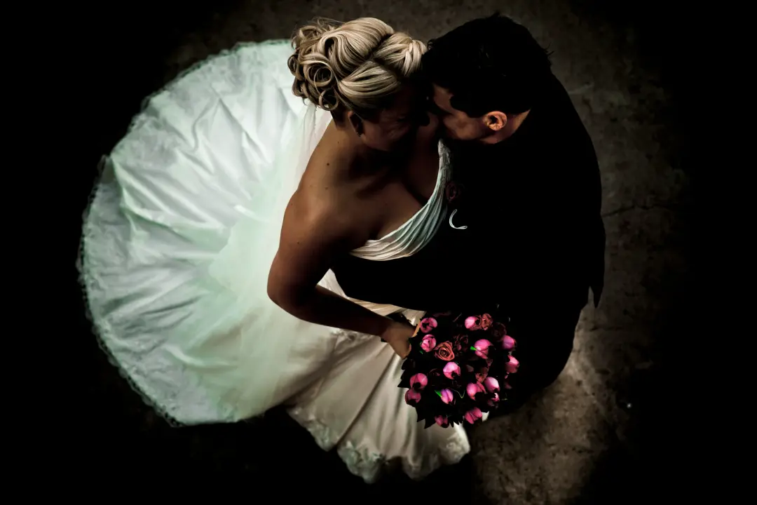 Bride and groom kissing photographed from above, her blonde hair pinned up over a pink rose bouquet