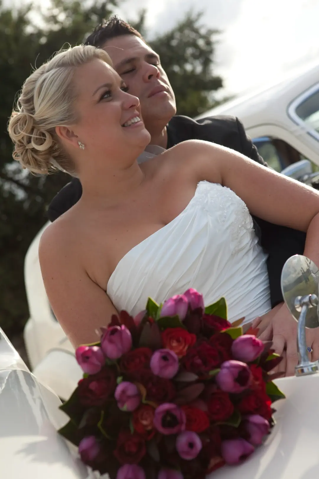 Bride with her groom in an open car, her blonde hair pinned low and a tulip bouquet in her lap