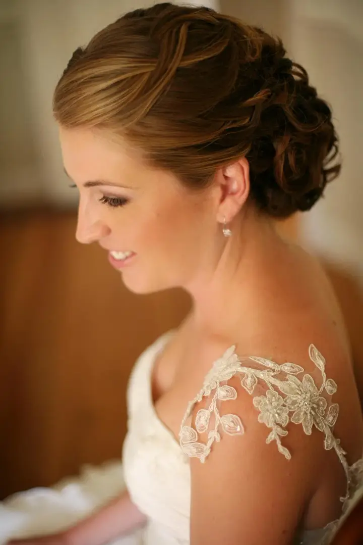 Bride in profile wearing a soft curled updo, her gown appliqued with lace at the shoulder