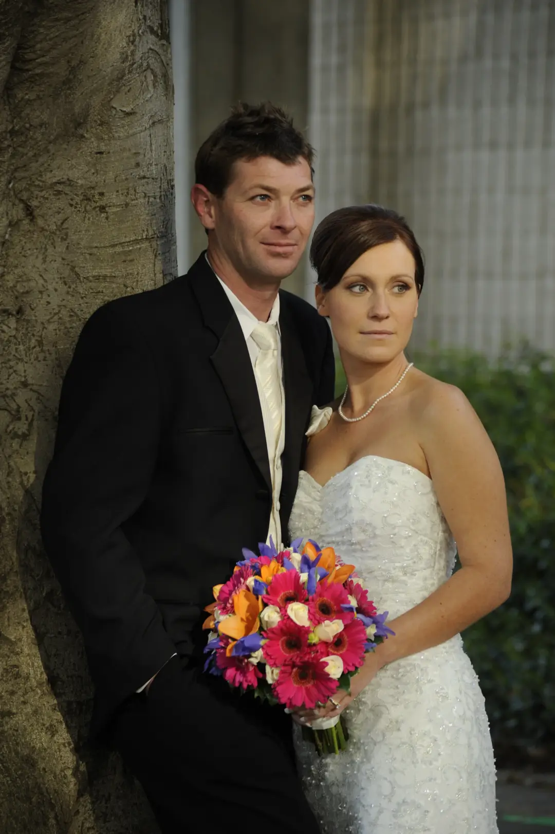Bride and groom against a tree, her dark hair pinned into a low style with a bright bouquet
