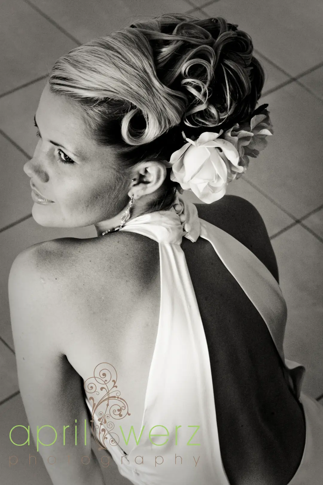Back view of a curled blonde upstyle dressed with a white rose, worn with a halterneck gown