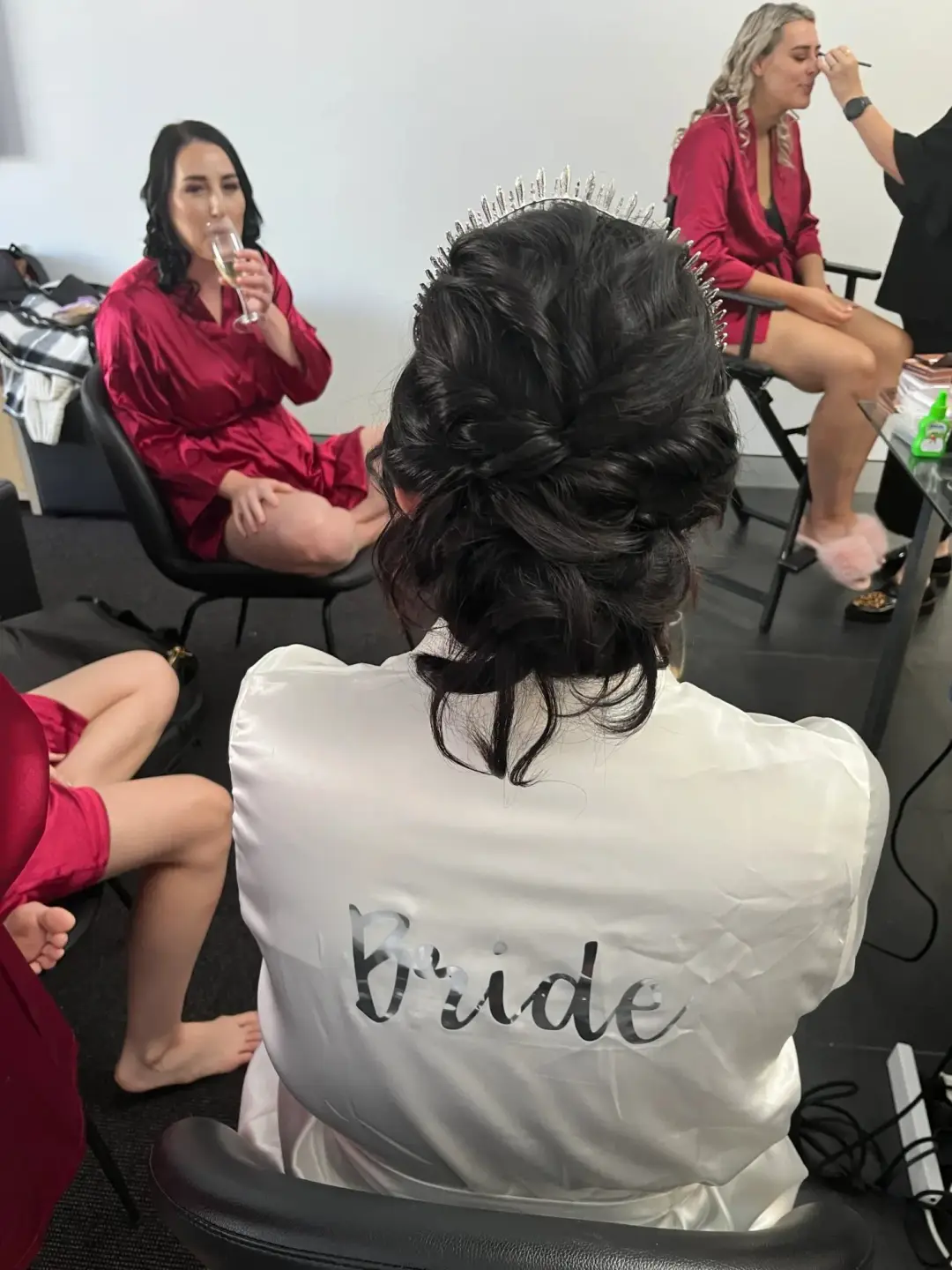 Bride in a white robe with a textured dark updo and a jewelled comb, her bridesmaids getting ready behind her