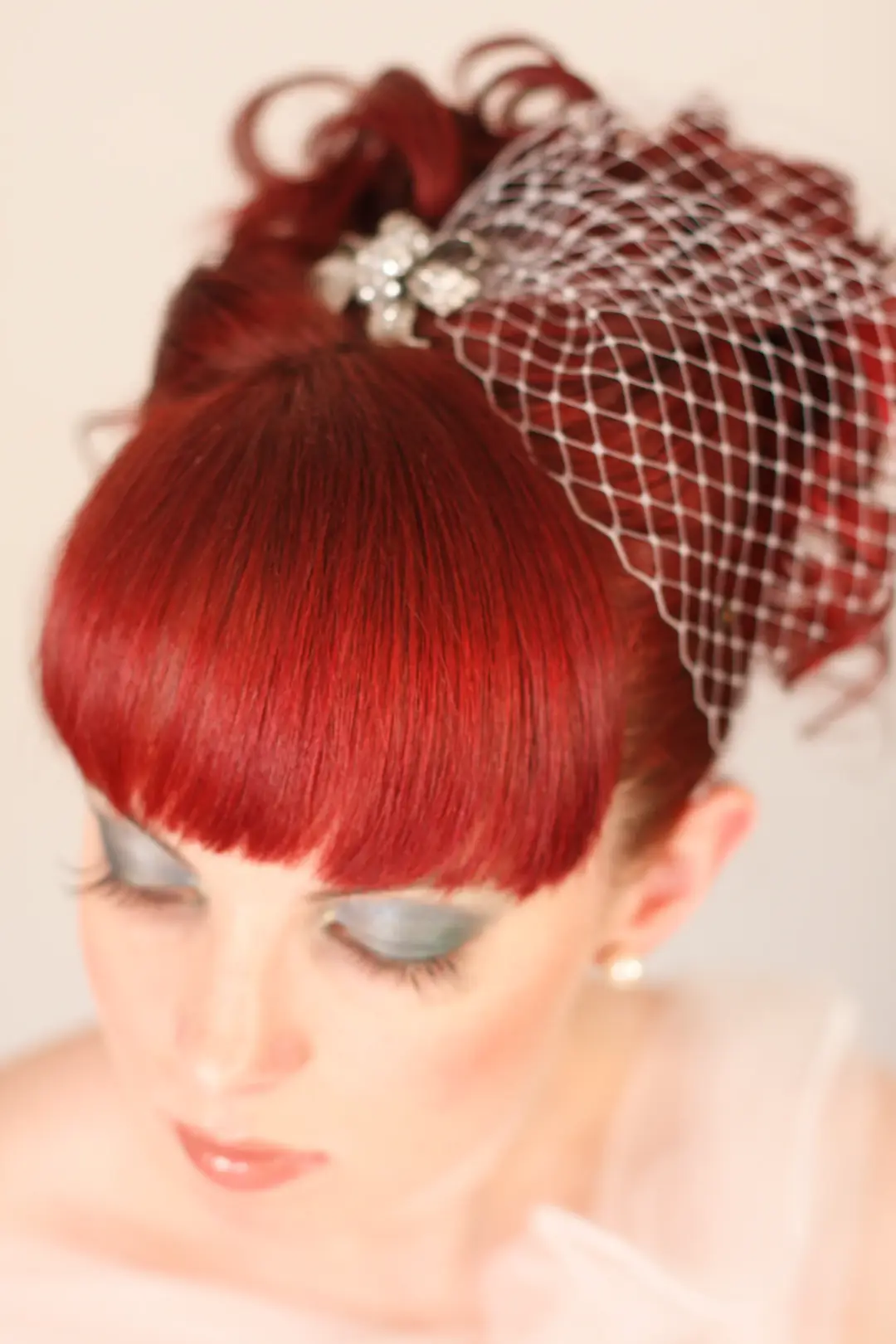 Red hair styled into a high curled updo with a blunt fringe, birdcage veil and silver eye makeup