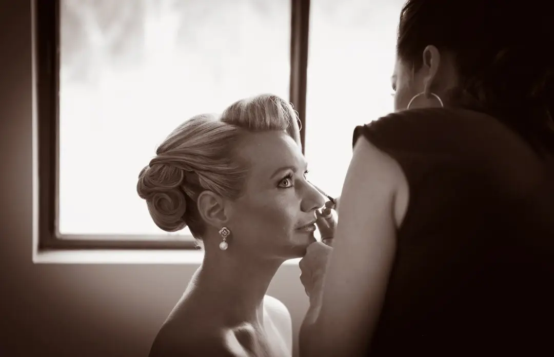 Makeup applied by the window on the wedding morning, the bride's blonde hair set in a French roll