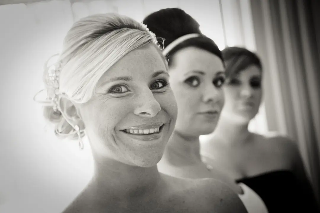 Bride smiling over her shoulder with her bridesmaids behind, her blonde hair pinned with a jewelled comb