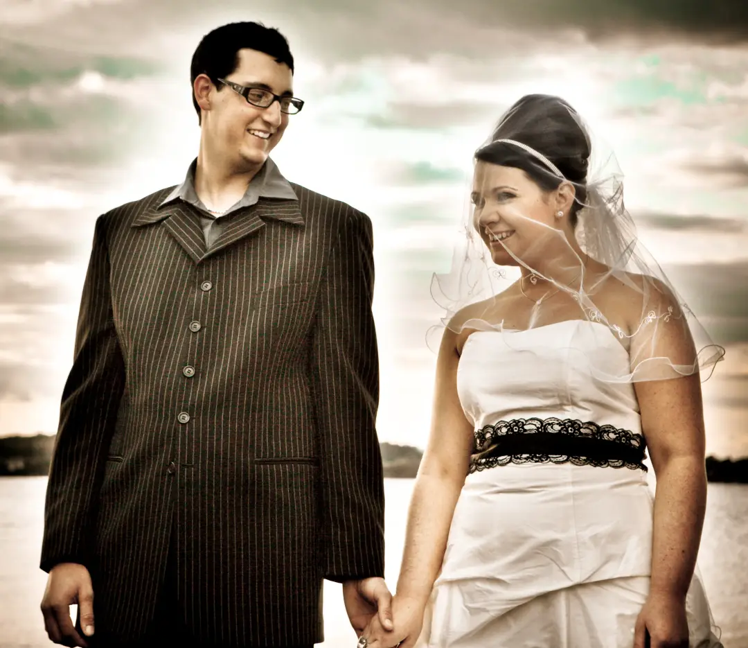 Bride and groom holding hands by the water, the bride's dark hair pinned up under a short veil