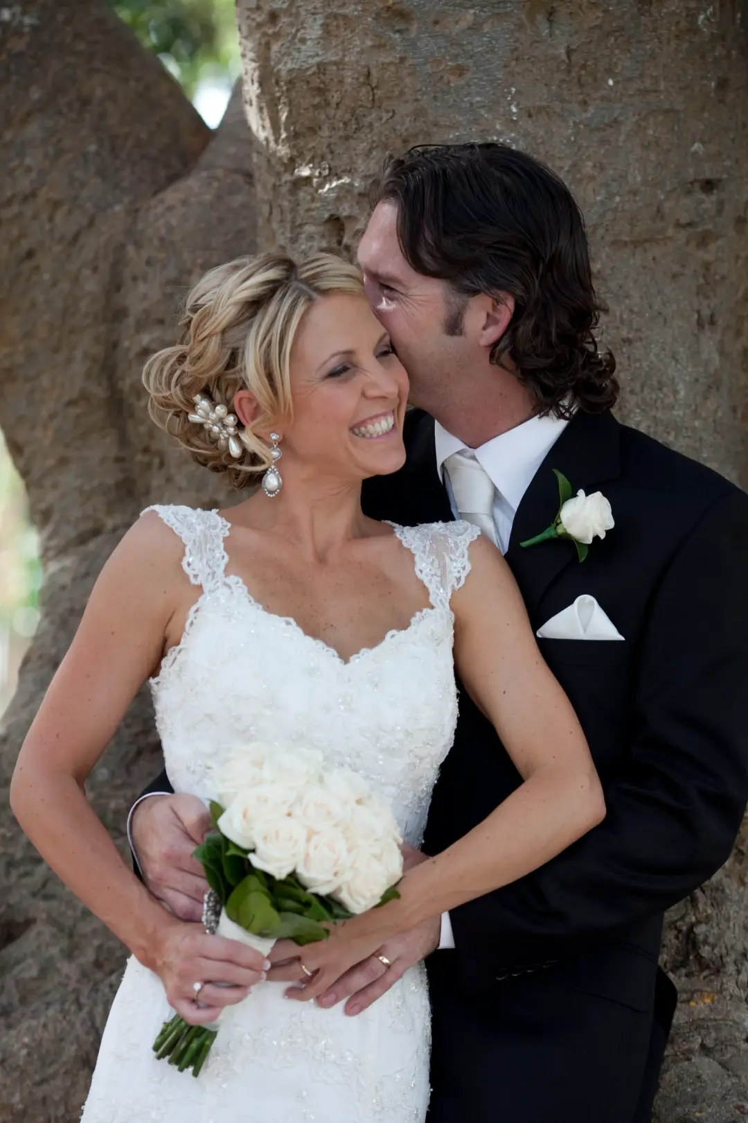 Newcastle bride and groom under a fig tree, the bride in a curled updo with a white floral hairpiece and a bouquet of white roses