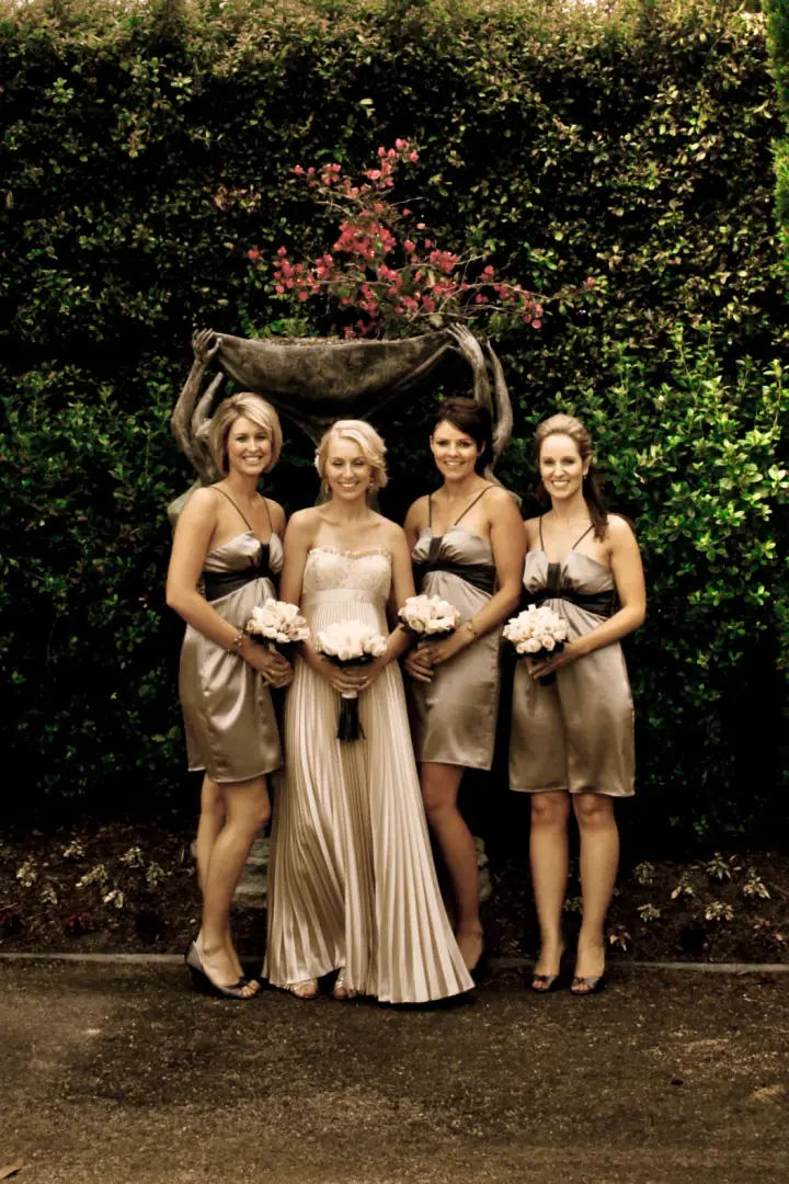 Bride and three bridesmaids in gold satin by a garden urn, all styled with pinned hair
