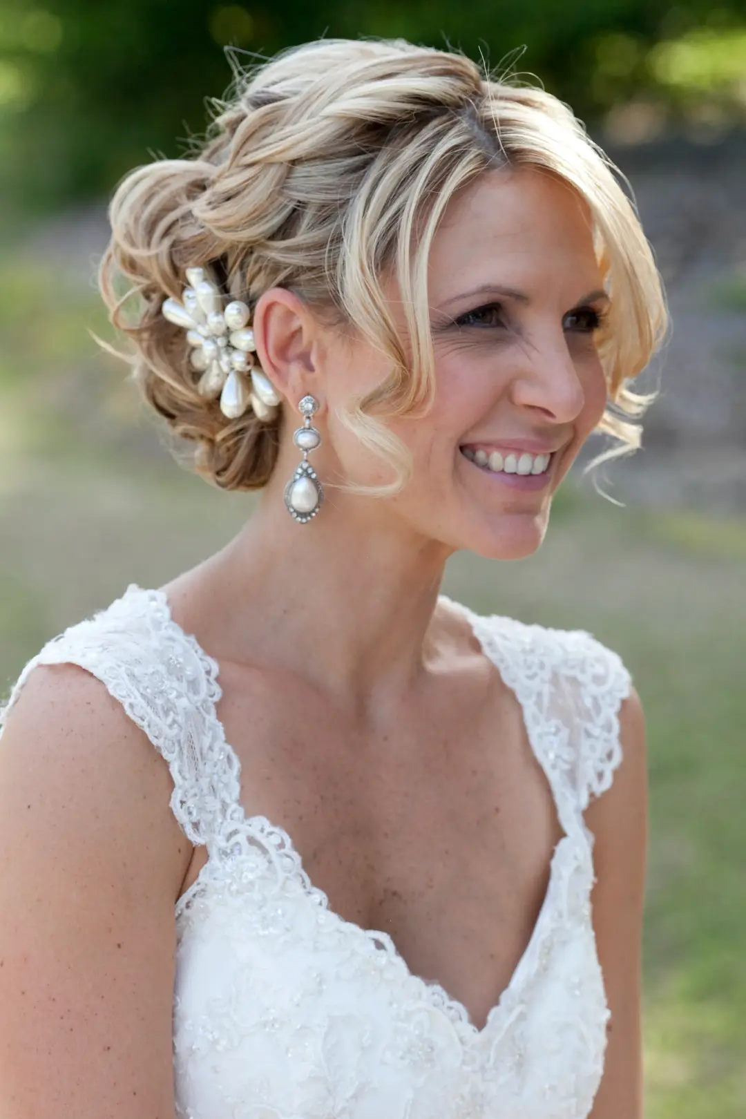 Blonde bride in a textured curled updo with a white flower hairpiece and pearl drop earrings