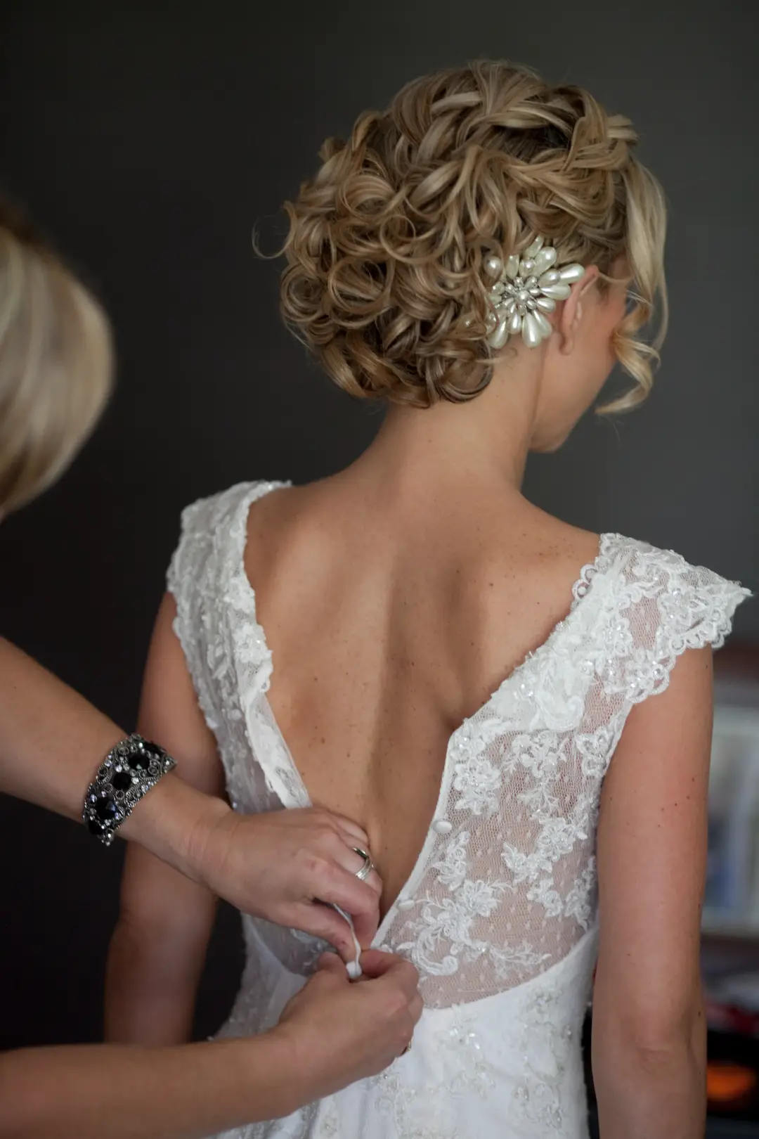Bride being buttoned into a lace gown, her blonde curled updo dressed with a jewelled comb