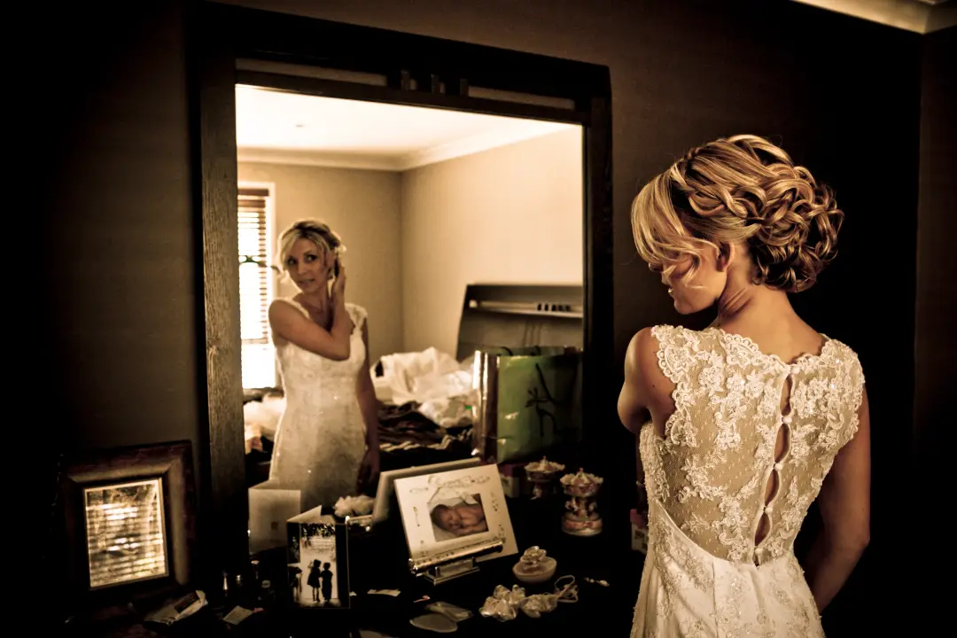 Bride at the mirror in a low curled updo, wearing a gown with a lace covered back