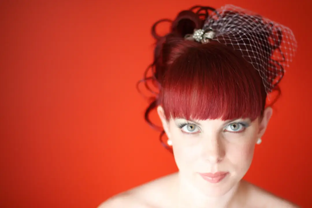 Formal portrait against red, a curled red updo with a birdcage veil and a jewelled clip