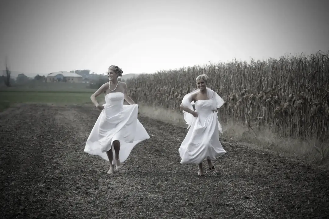 Two brides running along a farm track in white gowns, hair styled loose for the day, by Amanda Frost of Bellus