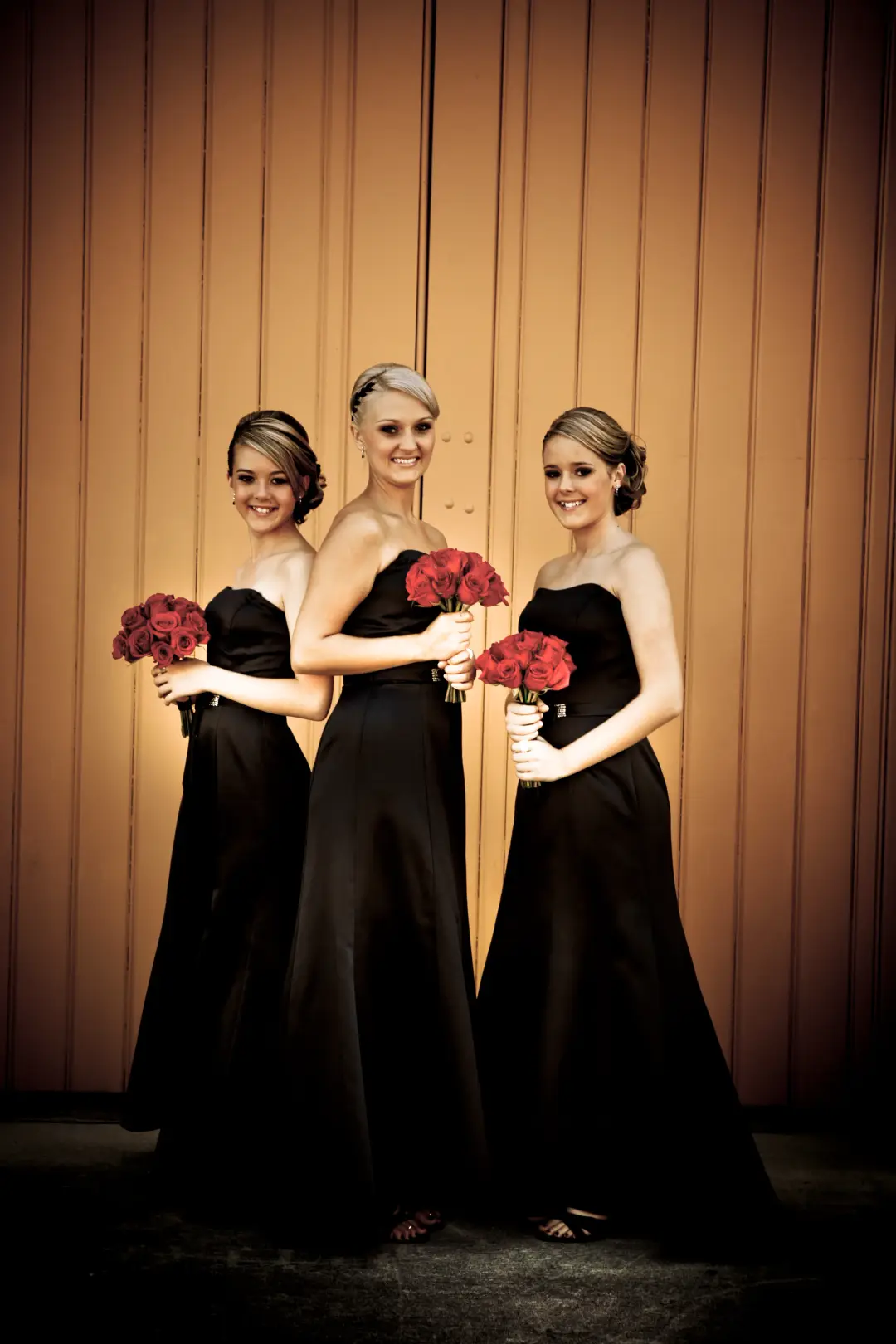 Bride and two bridesmaids in black with red rose bouquets, all three styled with pinned hair