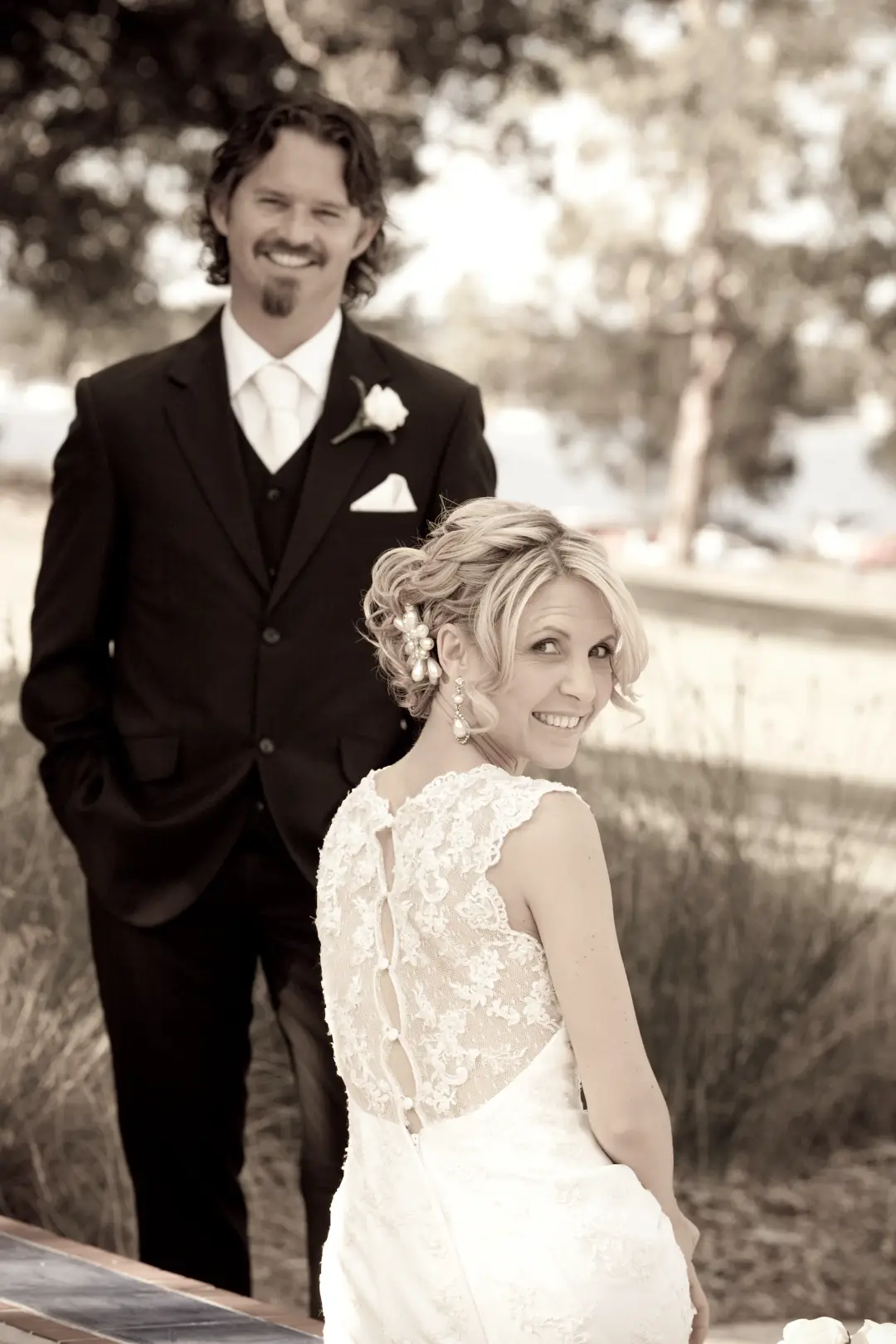 Bride glancing back at her groom, her blonde hair pinned with a small floral comb above a lace back
