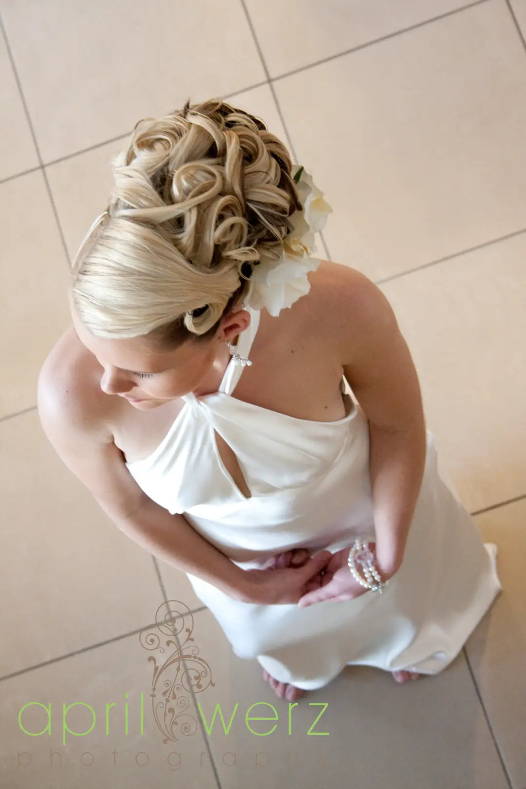 Curled blonde updo dressed with a white flower, photographed from above on a bride in a satin gown