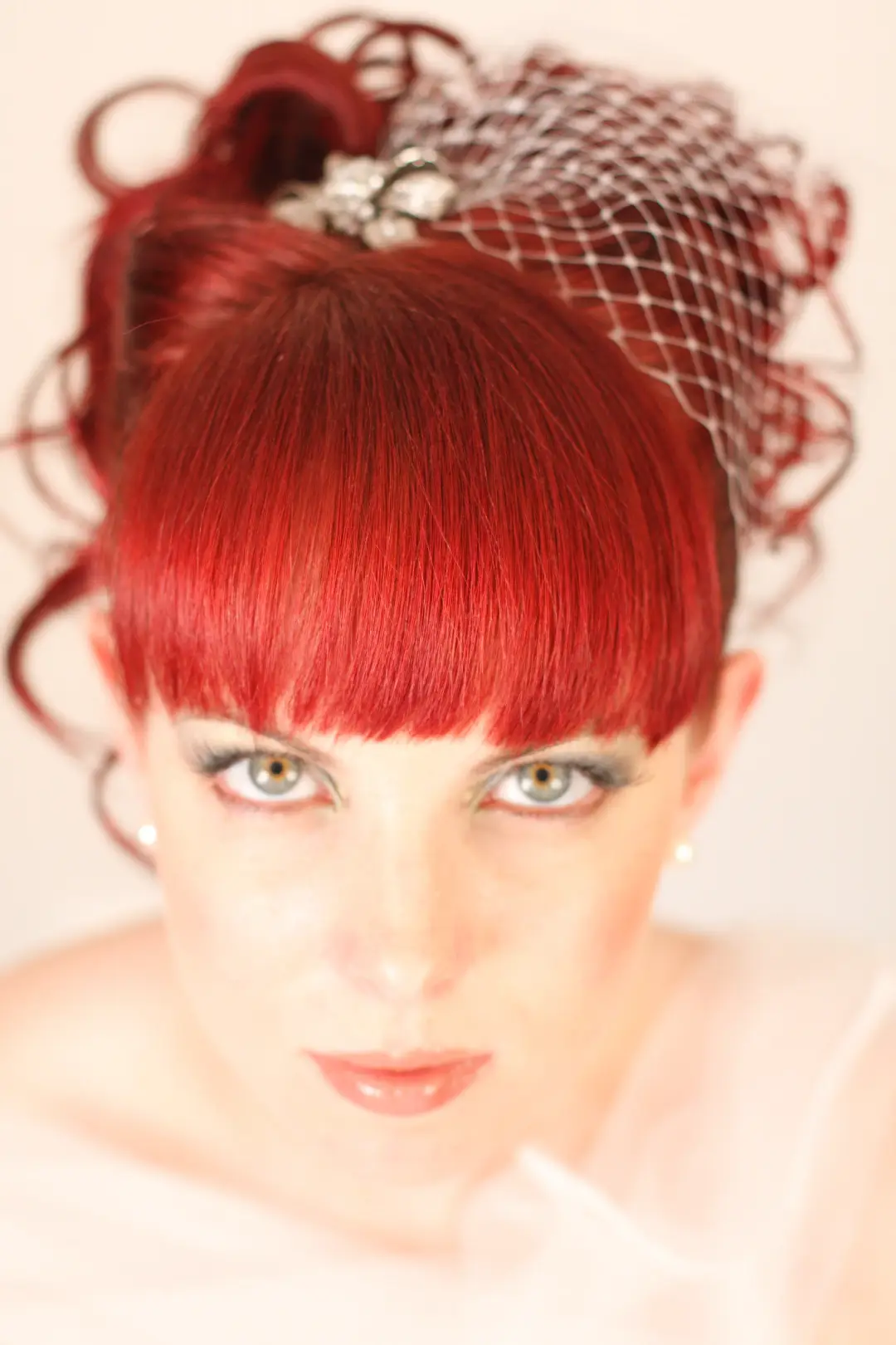 Red hair styled into a high curled updo with a blunt fringe and a birdcage veil