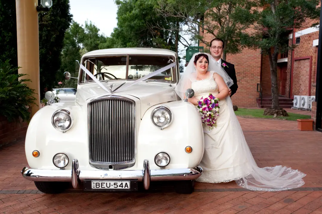 Bride beside a vintage wedding car, her hair pinned up with a cathedral veil