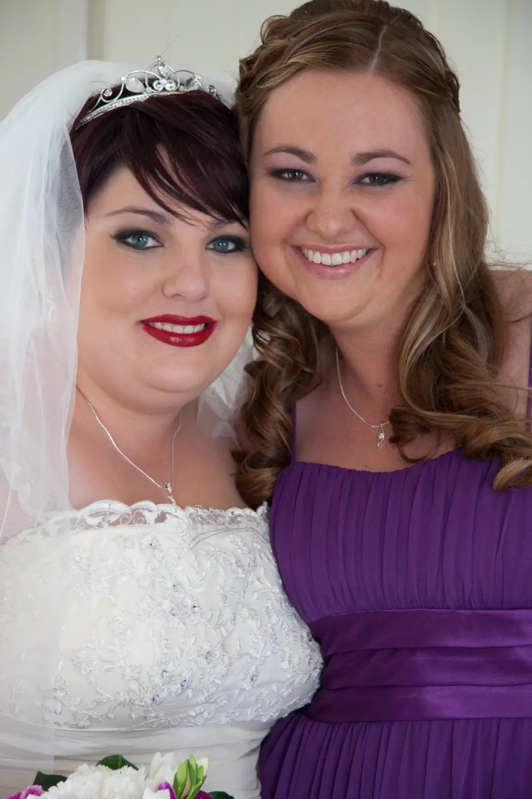 Bride in a tiara and veil with a bold red lip, beside a bridesmaid with long curled hair