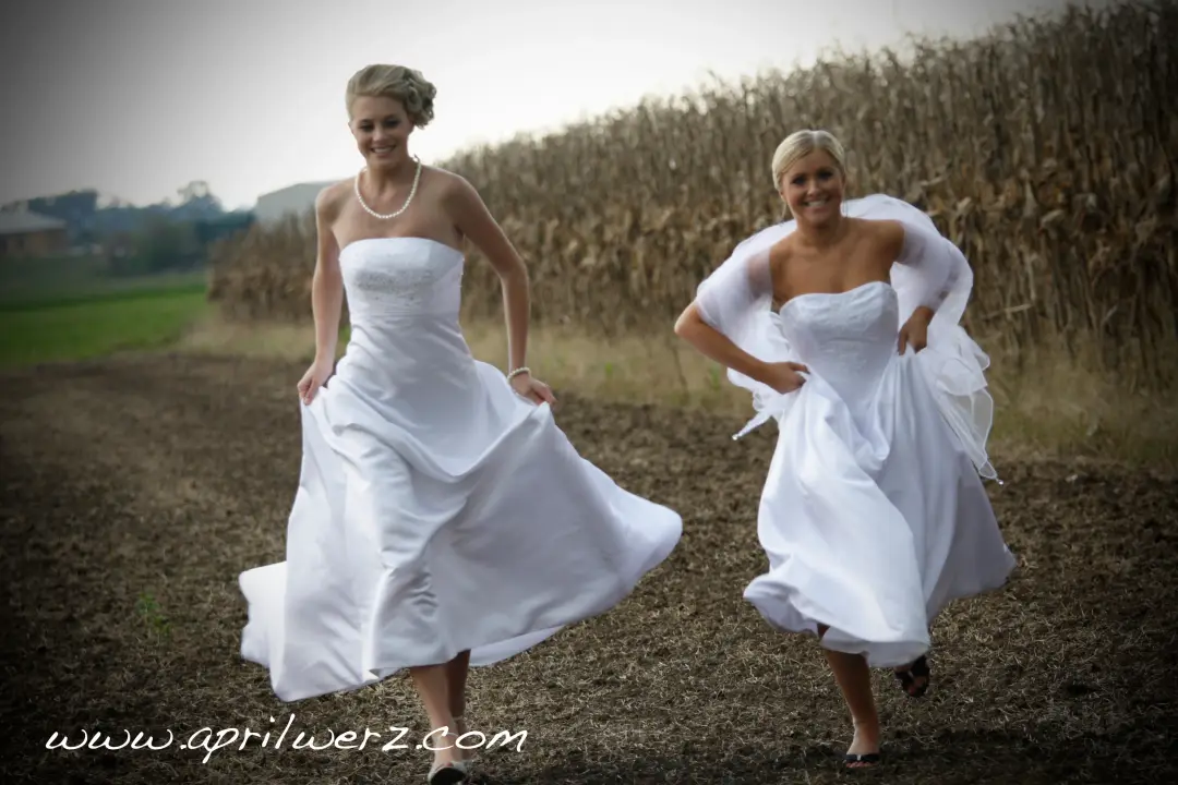 Two brides running along a farm track, one with a pinned updo and one with a veil
