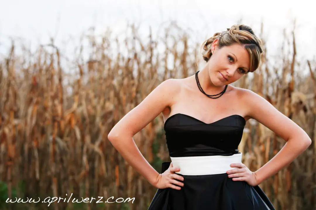 Guest in a black gown by a cornfield, her hair pinned into a curled updo