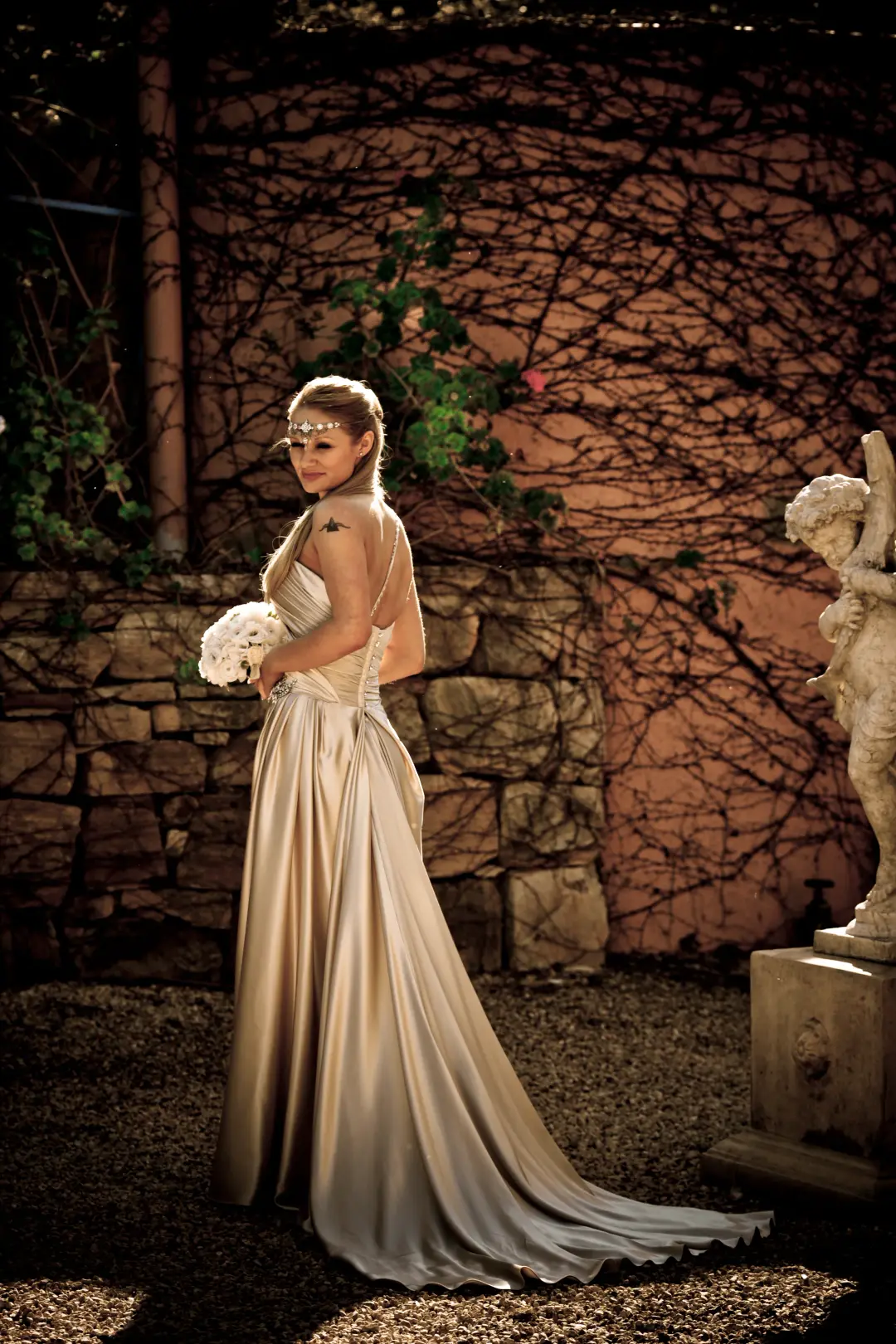 Bride in a satin gown against a stone wall, her hair pinned up with a jewelled headpiece across the brow