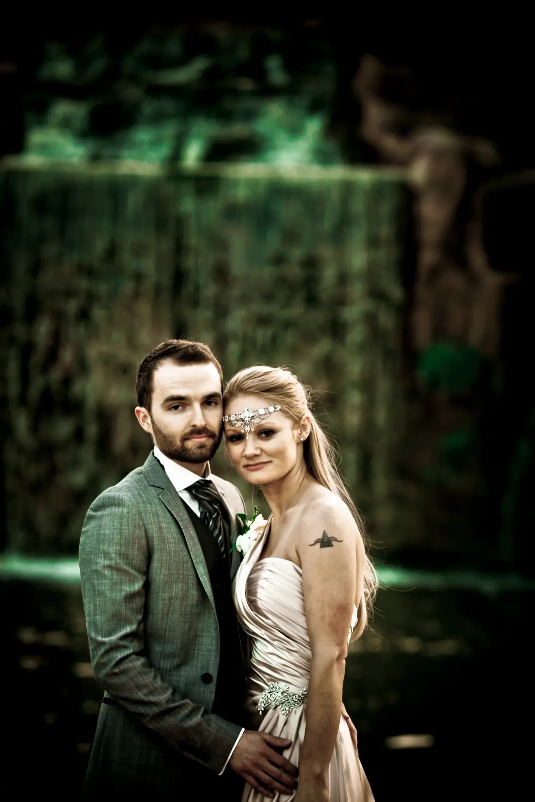 Bride and groom by a waterfall, her long blonde hair worn down with a jewelled forehead chain