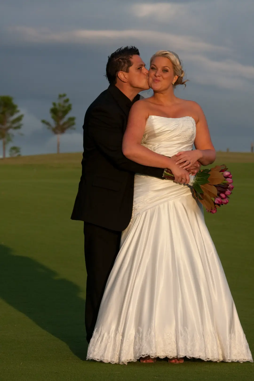 Groom kissing his bride on a golf course at dusk, her blonde hair pinned into a low updo