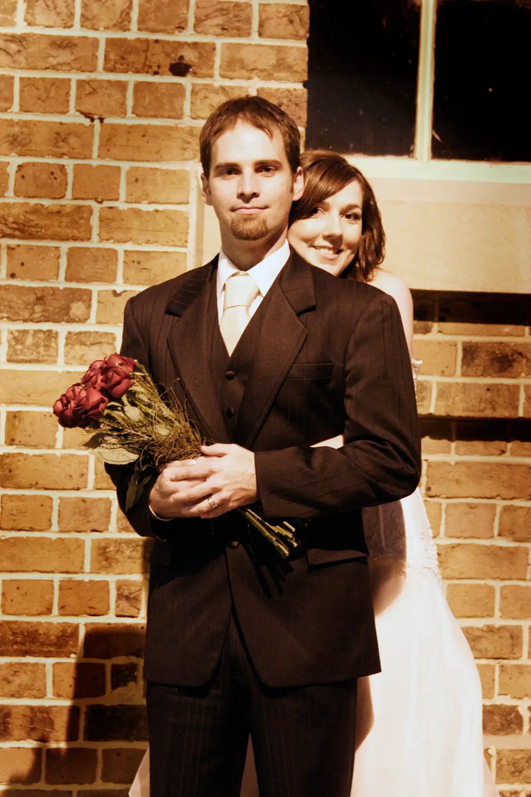 Bride and groom against a brick wall, the bride's dark hair pinned back under her veil