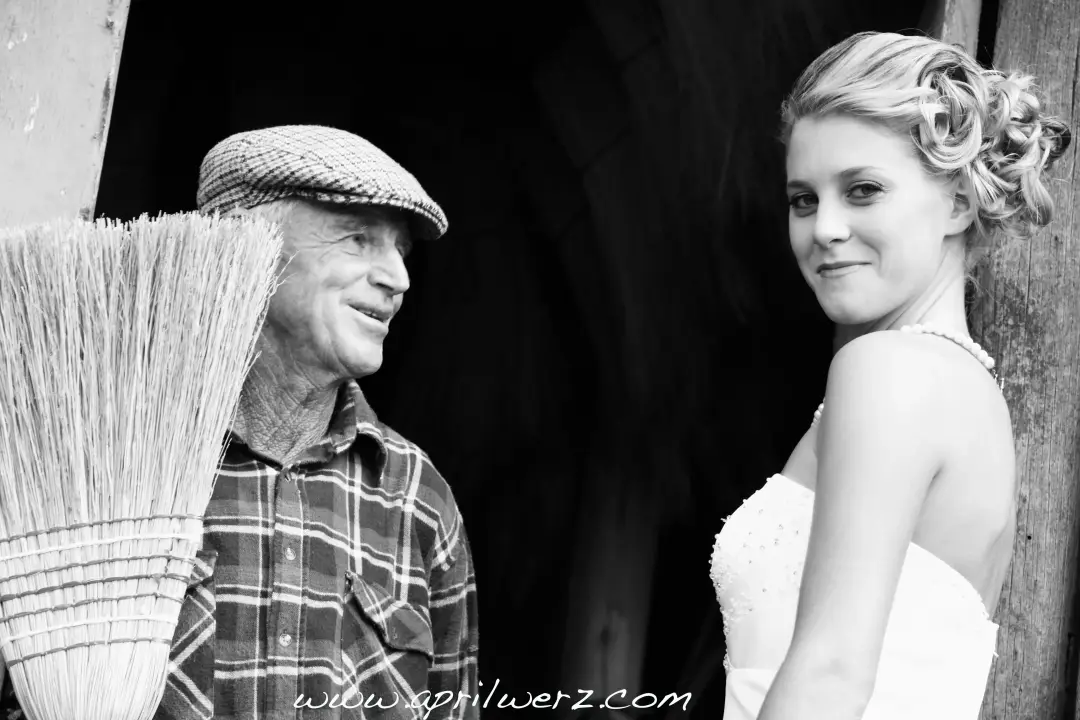 Bride talking with an older farmer in a barn doorway, her hair pinned up with a fabric flower