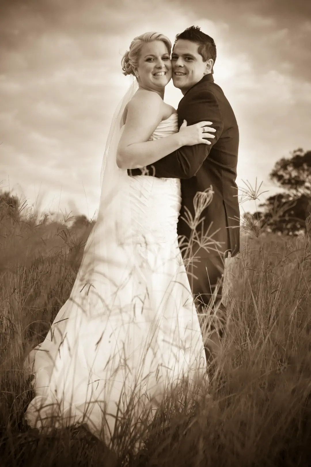 Bride and groom in long grass, her blonde hair pinned up beneath a full length veil