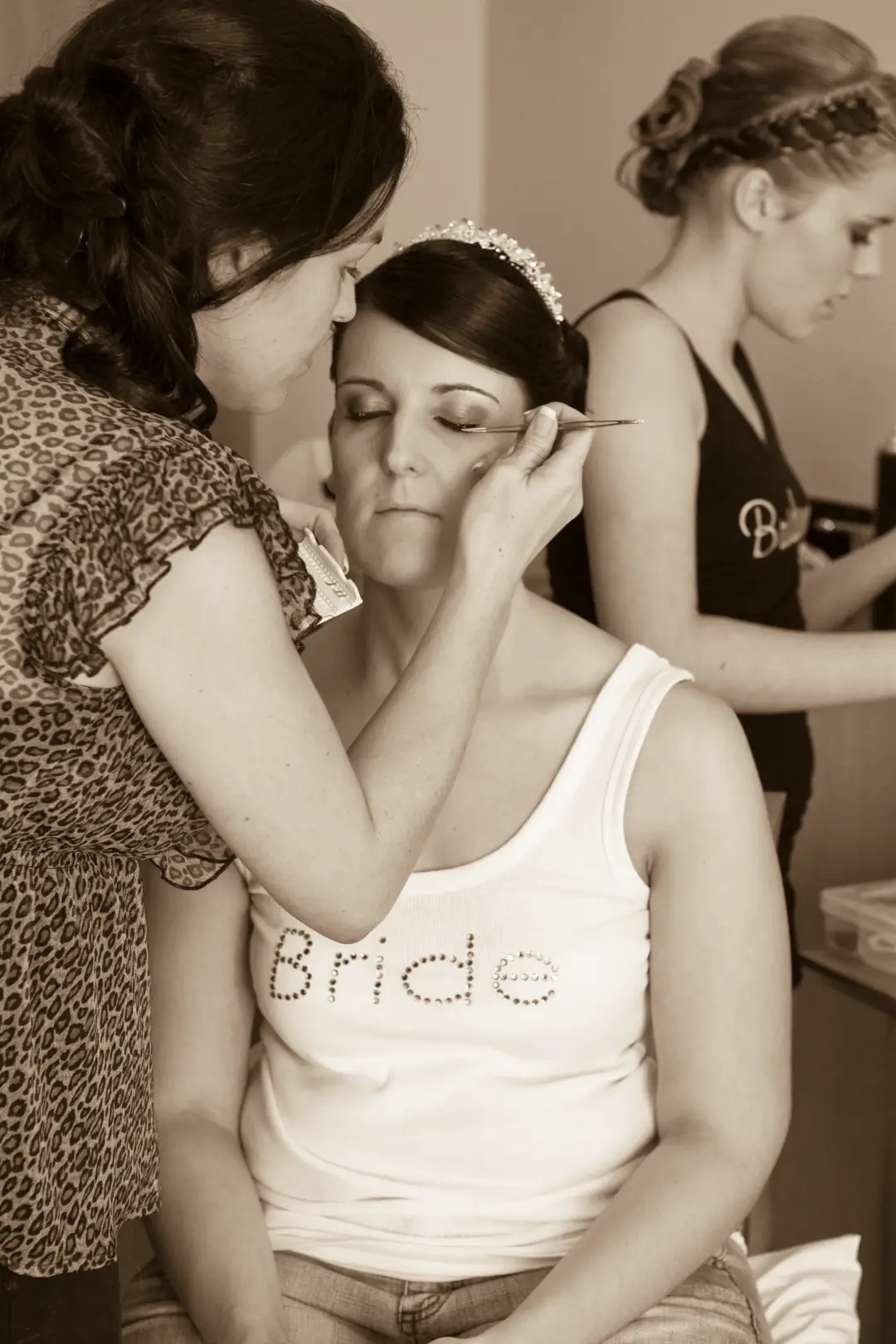 Bride having her eye makeup done in a bride singlet while a bridesmaid is styled behind her