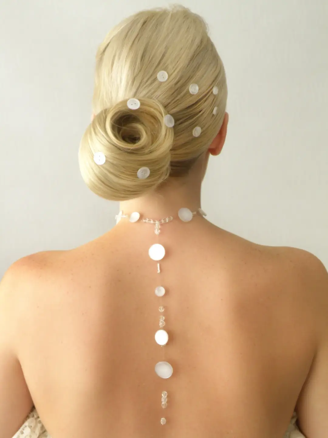 Back view of a rolled blonde bun set with pearl pins, worn with a backless gown and a draped back necklace