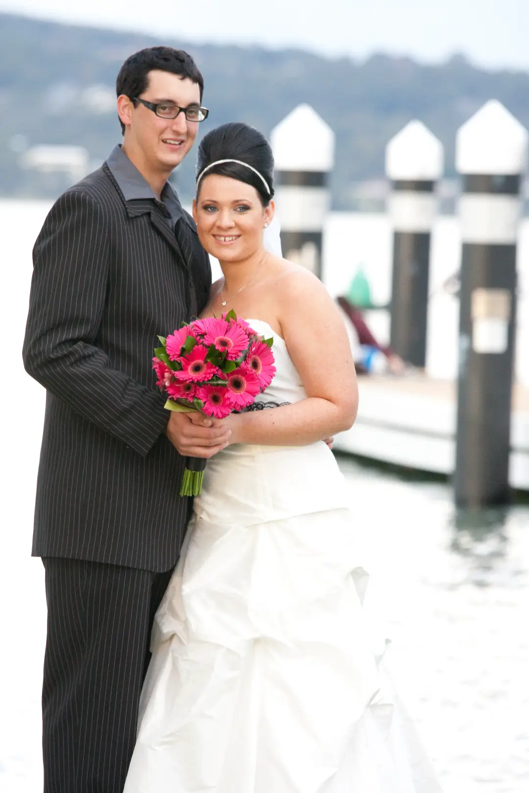 Bride and groom on the waterfront, the bride wearing a dark half up style with a headband and pink gerbera bouquet