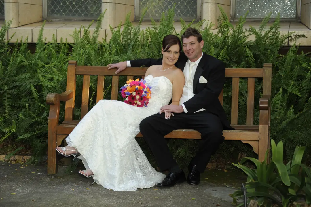 Bride and groom seated on a garden bench, the bride's dark hair pinned up with a bright bouquet in her lap