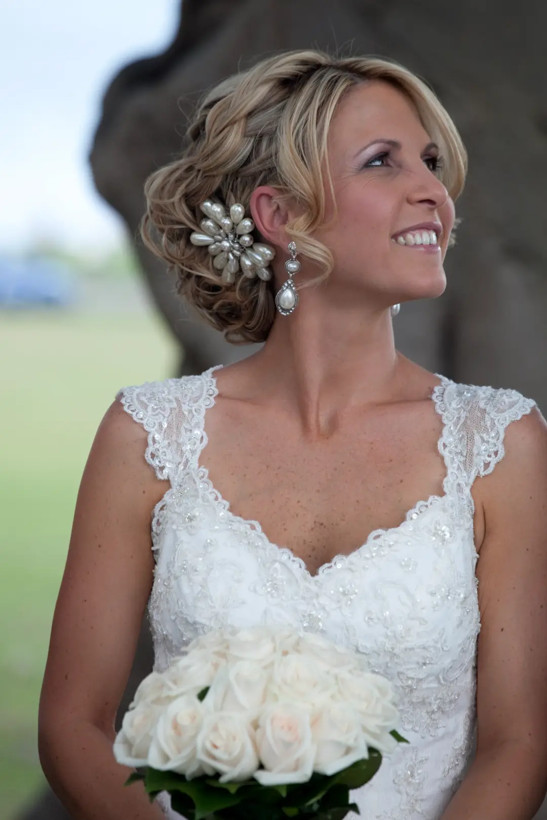 Bride in a curled blonde updo with a crystal hairpiece and pearl earrings, holding cream roses