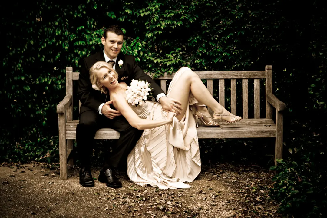 Bride lying across her groom's lap on a garden bench, her blonde hair pinned into a soft updo, by Amanda Frost of Bellus
