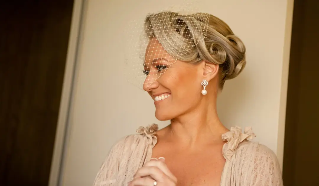 Bride in a birdcage veil over a blonde vintage roll, wearing warm natural makeup